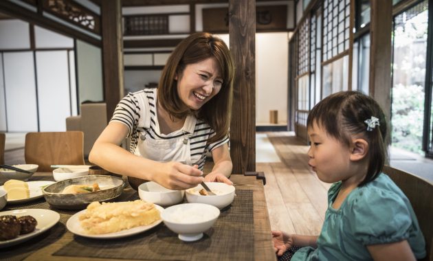 Young Japanese girl sitting at table with mother eating dinner, mother smiling, daughter watching